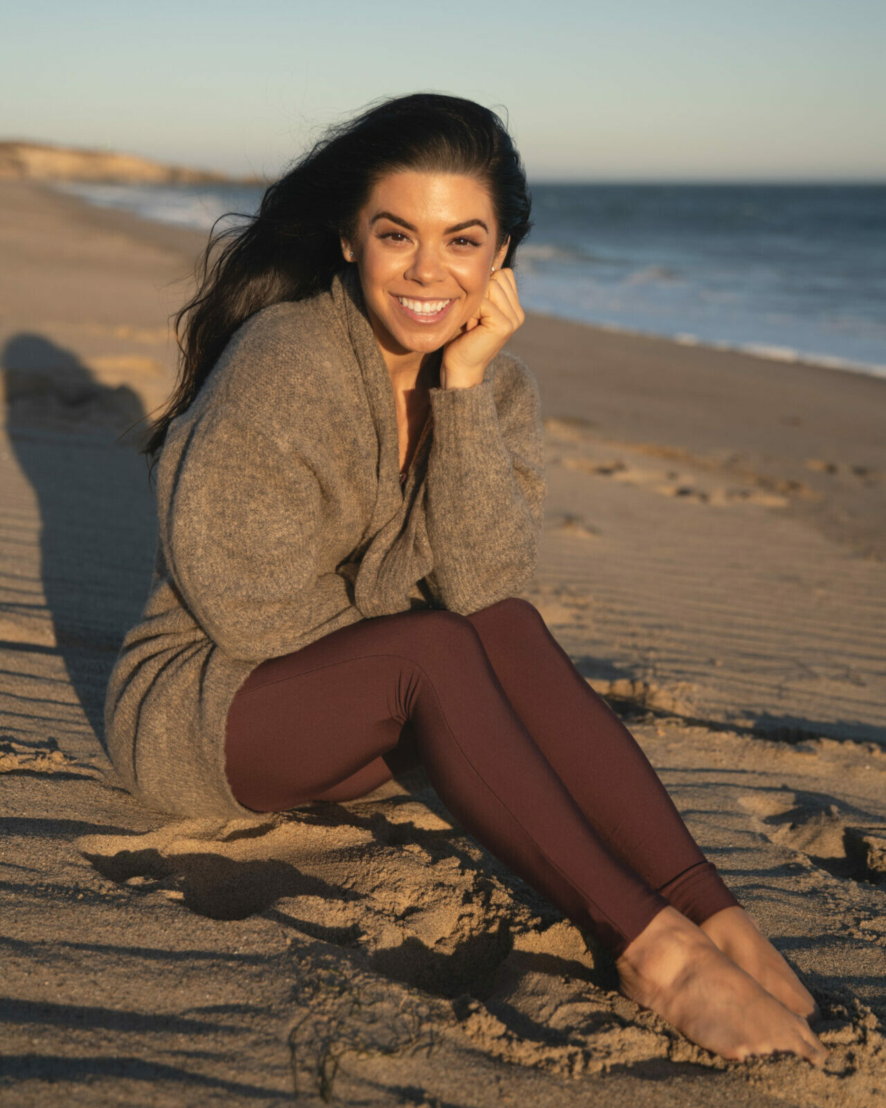 Gabby Sansosti sitting on the beach oceanside relaxed and joyful with a big smile during sunset