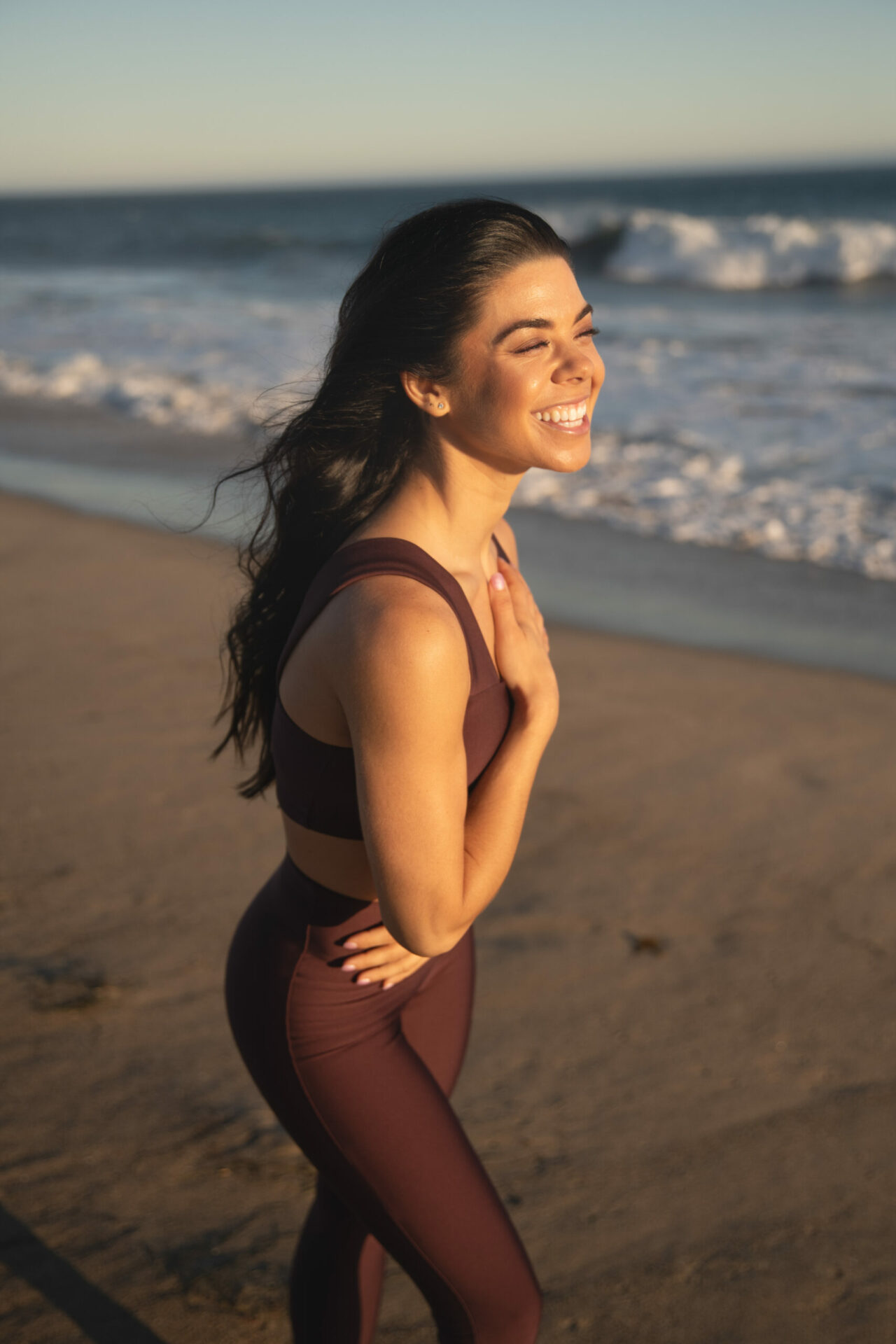 Gabby Sansosti in Malibu California on the beach next to the ocean in fitness set with her hand to her heart and a big smile full of joy as sunset is shining on her
